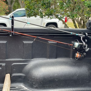 May include: Two fishing rods are secured in the back of a pickup truck bed. The rods are held in place by a black rod holder. The truck bed is lined with a black liner.
