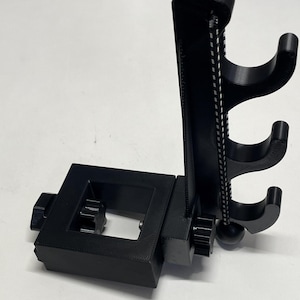 May include: A black 3D printed phone stand with a rotating arm and three hooks. The stand has a square base and a knob for adjusting the arm.