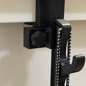 K&ouml;nnte beinhalten: Ein schwarzer, verstellbarer Klemmb&uuml;gel, der an einer horizontalen Fl&auml;che befestigt werden kann. Er verf&uuml;gt &uuml;ber einen sternf&ouml;rmigen Knopf zum Festziehen, ein schwarz-wei&szlig; gemustertes Bungee-Seil und mehrere Haken zum Aufh&auml;ngen von Gegenst&auml;nden. Der B&uuml;gel ist aus strapazierf&auml;higem Kunststoff gefertigt.