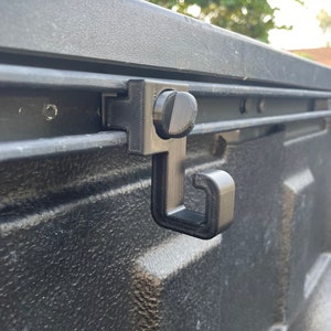 May include: A black plastic hook that attaches to a truck bed rail system. The hook is designed to hold items such as bags, ropes, or straps.
