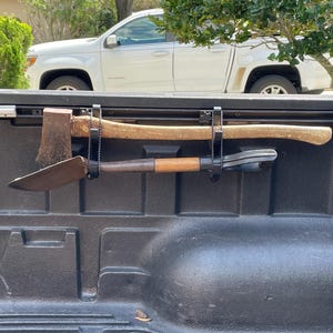 Peut inclure: Un plateau de camion noir avec un système de crémaillère en métal tenant une hache à manche en bois et une pelle. La hache et la pelle sont fixées avec des cordons élastiques noirs.