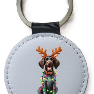 May include: A round keyring featuring a graphic of a dark-furred dog wearing brown reindeer antlers and a string of colourful Christmas lights wrapped around its body. The dog has a brown collar and is set against a light grey circular background.