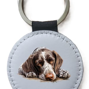 May include: A round keyring with a detailed illustration of a German Shorthaired Pointer dog. The dog is shown with brown and white markings, lying down with its eyes closed. The keyring has a silver ring and a grey leather-like backing.