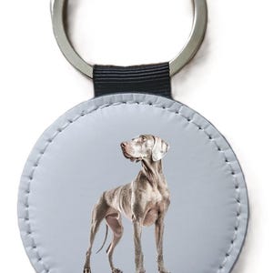 Peut inclure: Porte-clés rond avec une illustration de chien Weimaraner. Le chien est de couleur gris clair sur un fond bleu clair. Le porte-clés a un anneau en métal argenté et une sangle noire.