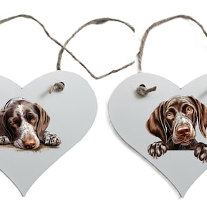 May include: Two heart-shaped wooden decorations featuring dog portraits. One shows a resting dog with brown and white fur, the other a dog with brown fur and paws. Both have twine hangers.