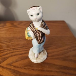 May include: A ceramic figurine of a cat-like figure, wearing a shawl and holding a basket. The figure has a white face with green eyes, a brown and black shawl, and a light blue dress. The figurine is standing on a small, round base.