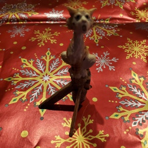 May include: A small figurine of a cat with orange and white fur, sitting on a wooden stand. The cat is looking up with its mouth open, as if it is meowing. The figurine is on a red background with a snowflake pattern.