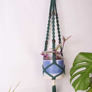 May include: A green macrame plant hanger with wooden beads holds a blue and white ceramic pot with a green plant.