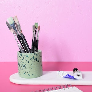 May include: A speckled green pot holds paintbrushes and a palette knife. Two paint tubes and a notebook sit on a white tray. The background is pink, with a plant in a white pot. The scene is set on a pink surface.