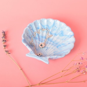 May include: A blue and white seashell-shaped dish with a silver chain necklace inside. The dish is on a pink background.