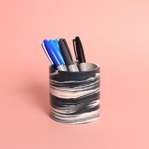 May include: A black and white marbled ceramic pencil holder with four pens inside. The pens are blue, grey, and black.
