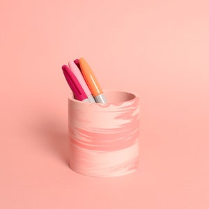 Könnte beinhalten: Ein rosa-weißer, marmorierter Stiftehalter aus Keramik mit drei Stiften darin. Die Stifte sind rosa, orange und grau.