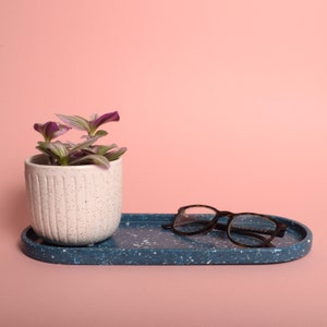 May include: A white ceramic planter with a textured surface, filled with a small purple and green plant, sits on a blue and white speckled oval tray with a pair of brown tortoiseshell glasses resting on the tray.