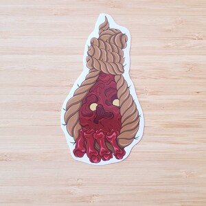 May include: A sticker featuring a stylized illustration of a red, fleshy face with yellow eyes and bone-like fingers. The face is framed by a brown rope, suggesting a noose. The sticker has a white border.