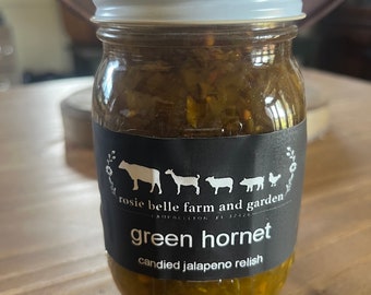 May include: A glass jar filled with green jalapeno relish. The jar has a black label with white text that reads "Rosie Belle Farm and Garden, Candleton, FL 32120, Green Hornet, Candied Jalapeno Relish". The label also features a silhouette of a cow, goat, pig, and chicken.