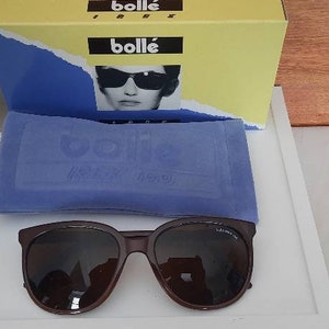 May include: Brown sunglasses with a dark lens and a blue velvet case. The case has the word "Bolle" printed on it. The sunglasses are in a box with the word "Bolle" printed on it.