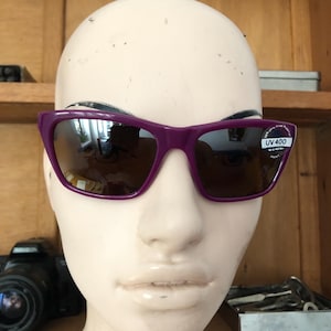 May include: Purple sunglasses with dark tinted lenses and a rectangular frame shape. A small circular sticker on the right lens displays the text UV 400 and indicates 100% Ultra Violet Protection. The glasses are positioned on a neutral mannequin head.