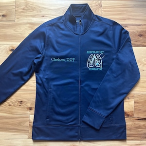 May include: A navy blue zippered jacket with the text "Chelsea, RRT" and "Respiratory Therapist" embroidered on the front. The embroidery also includes a stylized image of lungs.
