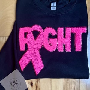 May include: Black sweatshirt with a pink ribbon and the word "FIGHT" in pink glitter lettering. The sweatshirt is a popular item for breast cancer awareness.