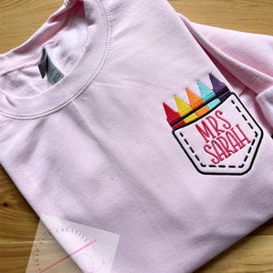 May include: Pink long-sleeved shirt with a rainbow crayon pocket and the text "Mrs. Sarah" embroidered on it.
