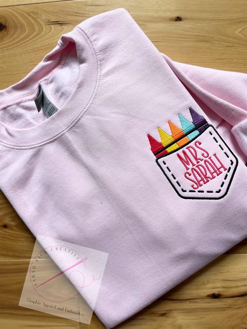 May include: Pink long-sleeved shirt with a rainbow crayon pocket and the text "Mrs. Sarah" embroidered on it.
