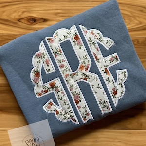 May include: A light blue sweatshirt with a white scalloped monogram featuring the letters "R.F." in a floral print. The floral print features small white flowers with orange and pink accents.