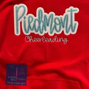 May include: Red hooded sweatshirt with a white and teal blue embroidered design that says "Piedmont Cheerleading".