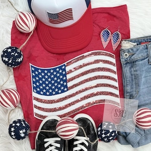 May include: Red, white, and blue themed outfit featuring a red t-shirt with a sequined American flag design, a white baseball cap with a red brim and an American flag patch, blue denim jeans, and red, white, and blue striped and star-shaped ornaments.