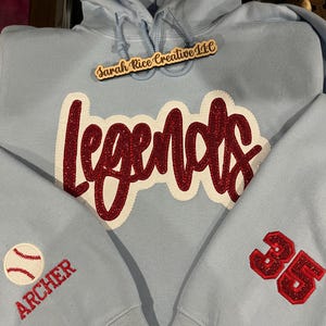May include: Light blue hoodie with a red glitter "Legends" design on the front, a baseball with the word "Archer" on one sleeve, and the number "35" on the other sleeve. The hoodie also has a wooden tag that says "Sarah Rice Creative 11C".