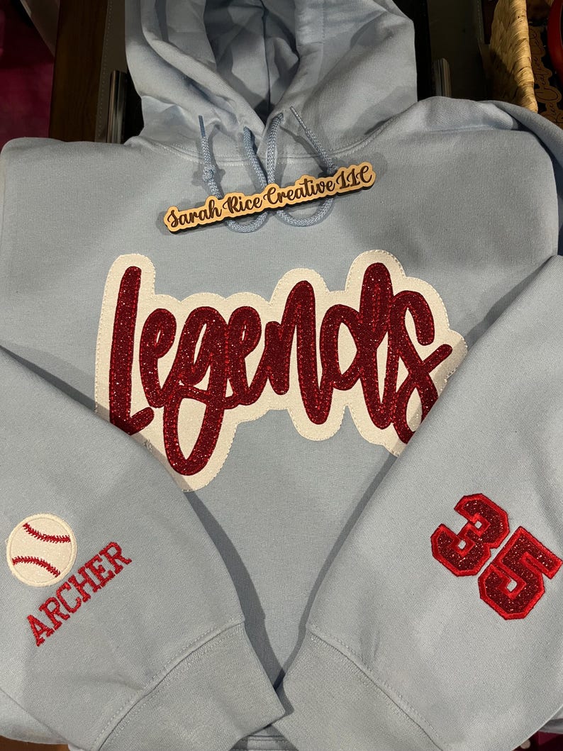 May include: Light blue hoodie with a red glitter "Legends" design on the front, a baseball with the word "Archer" on one sleeve, and the number "35" on the other sleeve. The hoodie also has a wooden tag that says "Sarah Rice Creative 11C".