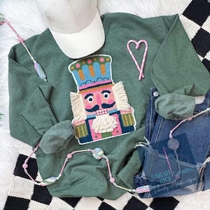 May include: An olive green sweatshirt with a colorful nutcracker applique, a white corduroy baseball cap, and a pair of blue jeans. A pink and white candy cane heart and a beaded necklace are also visible. The sweatshirt is part of a holiday collection.