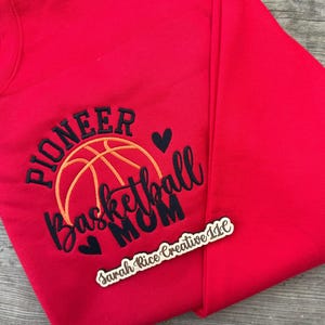 Pu&ograve; includere: Una felpa rossa con la scritta ricamata "PIONEER Basketball MOM" in nero e arancione, con un disegno di pallone da basket e due cuori neri. Un piccolo cartello di legno con testo &egrave; in basso a destra.