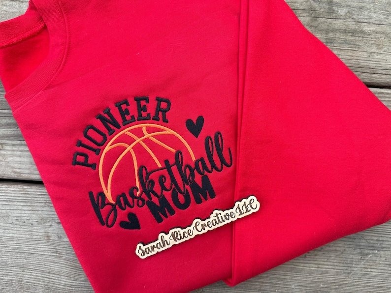 Pu&ograve; includere: Una felpa rossa con la scritta ricamata "PIONEER Basketball MOM" in nero e arancione, con un disegno di pallone da basket e due cuori neri. Un piccolo cartello di legno con testo &egrave; in basso a destra.