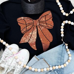 May include: Black sweatshirt with a large copper-colored sequined bow. The sweatshirt is laying on a pair of blue jeans and a string of wooden beads.