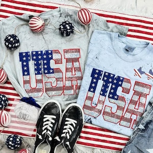May include: Two light blue t-shirts with the word "USA" in red, white, and blue glitter letters. The shirts are on a red and white striped background with red, white, and blue decorative stars.