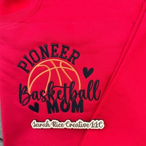 Pu&ograve; includere: Una felpa rossa con la scritta ricamata "Pioneer Basketball Mom" in nero e oro. Un disegno di pallacanestro &egrave; sopra il testo, con due piccoli cuori neri. Il testo "Sarah Rice Creative LLC" &egrave; in basso.