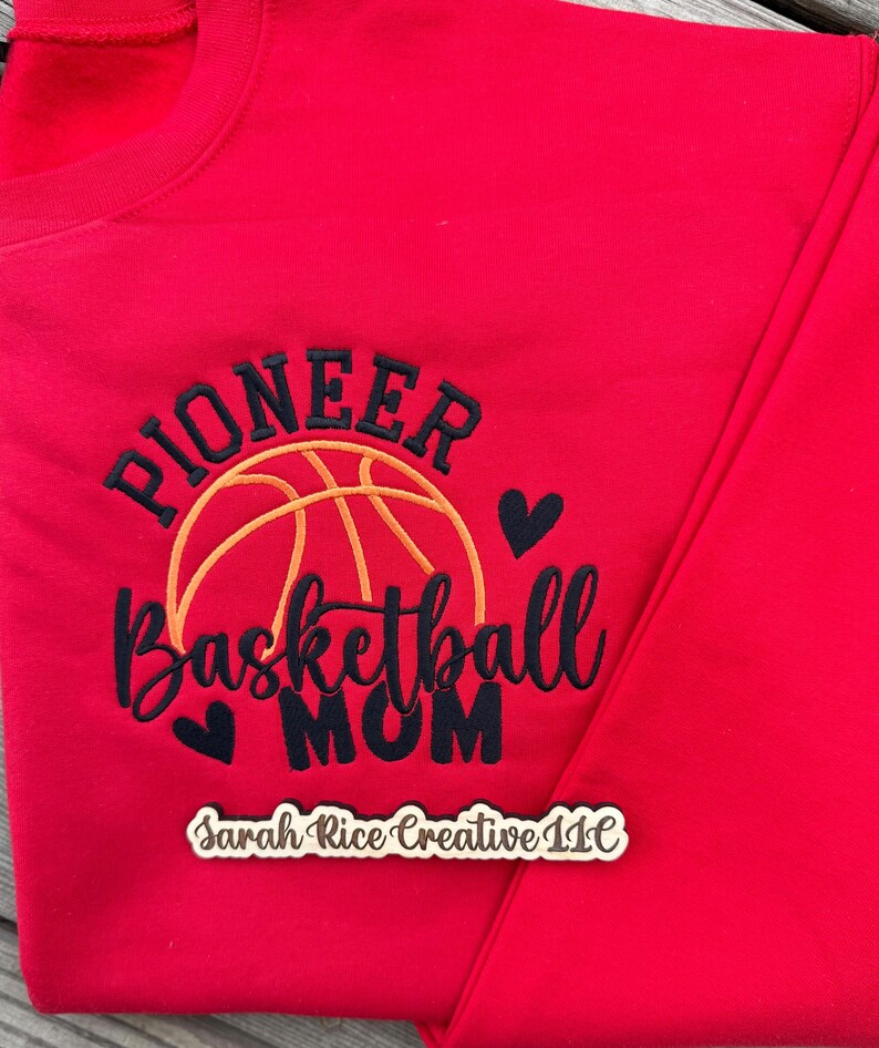 Pu&ograve; includere: Una felpa rossa con la scritta ricamata "Pioneer Basketball Mom" in nero e oro. Un disegno di pallacanestro &egrave; sopra il testo, con due piccoli cuori neri. Il testo "Sarah Rice Creative LLC" &egrave; in basso.