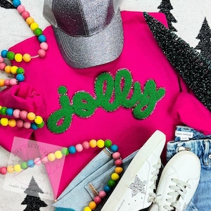 May include: A pink sweatshirt with a green felt "Jolly" applique. The sweatshirt is styled with a silver glitter baseball cap, a rainbow beaded necklace, and white sneakers with glitter stars.