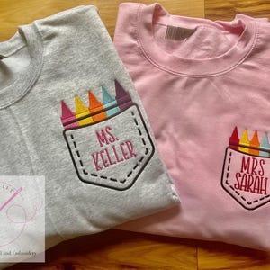 May include: Two gray and pink t-shirts with embroidered crayon pocket designs. The gray shirt says "Ms. Keller" and the pink shirt says "Mrs. Sarah".