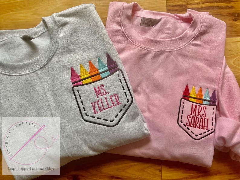 May include: Two gray and pink t-shirts with embroidered crayon pocket designs. The gray shirt says "Ms. Keller" and the pink shirt says "Mrs. Sarah".