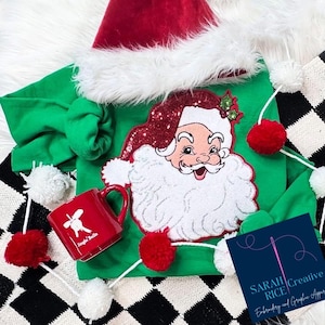May include: A green Christmas outfit with a Santa Claus graphic, a red Santa hat, and a red mug that says "Jingle Bells". The outfit is decorated with red and white pom-poms. The image also includes a black and white checkered pattern.