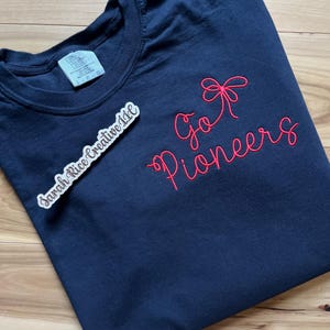 May include: Navy blue t-shirt with red embroidered text that reads "Go Pioneers" with a bow design. The shirt has a crew neck and a tag that reads "Essential Colors". A sticker with the text "Sarah Rice Creative LLC" is also visible.