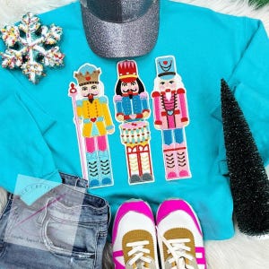 May include: A turquoise blue sweatshirt with three embroidered nutcracker figures. The nutcrackers are wearing colorful hats and outfits. The sweatshirt is laid out on a white surface with a pair of brown and pink sneakers and a pair of blue jeans.