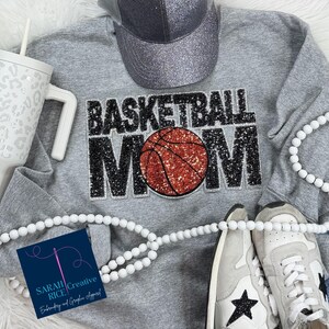 May include: Gray sweatshirt with the words "Basketball Mom" in black glitter with a red and orange basketball in the center. The sweatshirt is on a white surface with white beads and a gray and silver baseball cap.