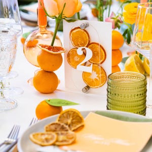 Puede incluir: Una tarjeta de número de mesa blanca con el número "2" impreso en blanco sobre un fondo acrílico transparente. La tarjeta está decorada con rodajas de naranja secas. La mesa está puesta con un mantel blanco, fruta naranja y cristalería verde.