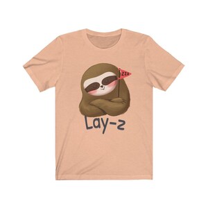 May include: A peach-colored t-shirt with a cartoon sloth holding a red flag with the text "zzz" on it. The text "Lay-z" is printed below the sloth.
