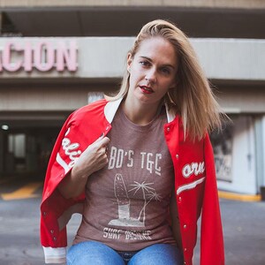 May include: A woman wearing a red bomber jacket with white lettering, a brown t-shirt with the text "BO'S TGC SURF DESIGNS", and blue jeans.
