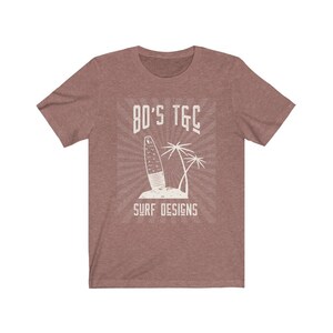 May include: A heather brown t-shirt with a vintage-style graphic of a surfboard and palm trees. The text "80's TGC Surf Designs" is printed on the shirt.