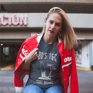 May include: A woman wearing a red varsity jacket with white lettering, a grey t-shirt with a graphic of a surfboard and palm trees, and blue jeans. The t-shirt has the text "80's TGC Surf Designs" printed on it.