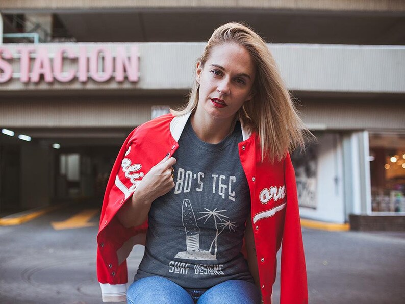 May include: A woman wearing a red varsity jacket with white lettering, a grey t-shirt with a graphic of a surfboard and palm trees, and blue jeans. The t-shirt has the text "80's TGC Surf Designs" printed on it.